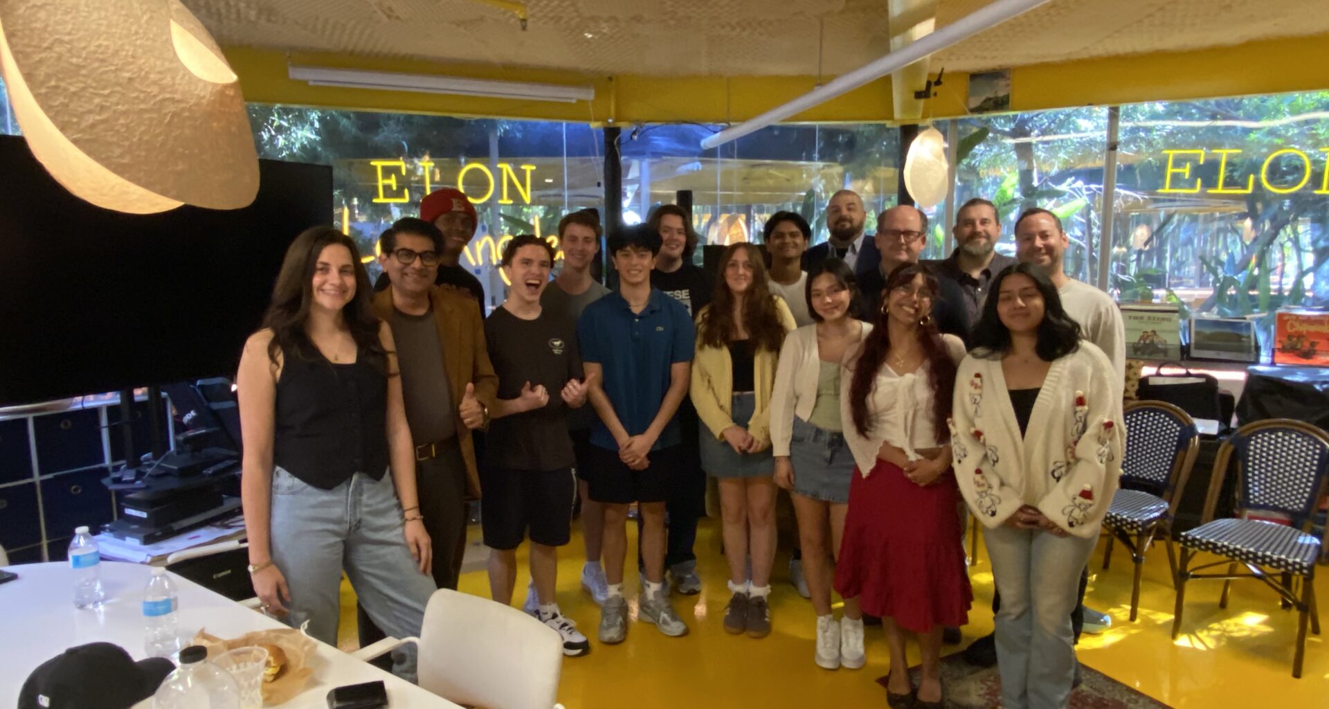 A group of students and staff stand together smiling in a bright room during an Elon University Los Angeles orientation event. Tables, chairs, and windows with “ELON” signage are visible, creating a welcoming group setting.