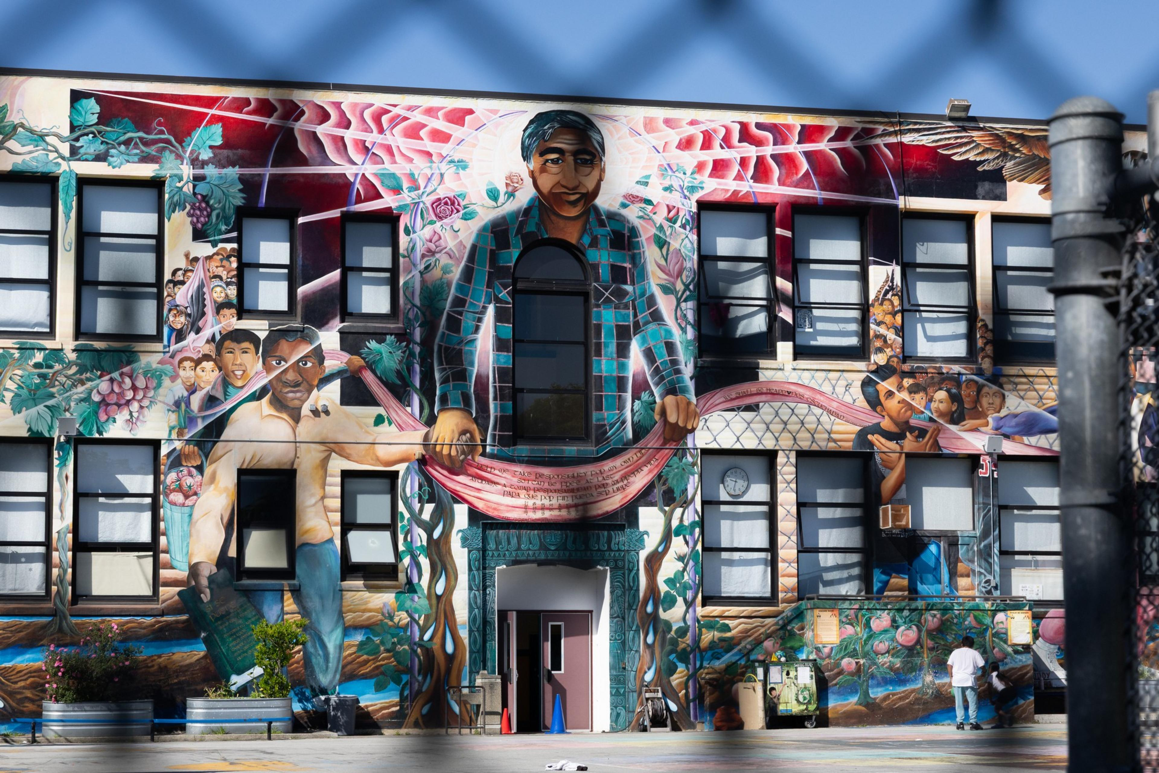 A vibrant mural spans a building, depicting a central figure holding a pink banner, with scenes of people, grapes, and nature surrounding him.