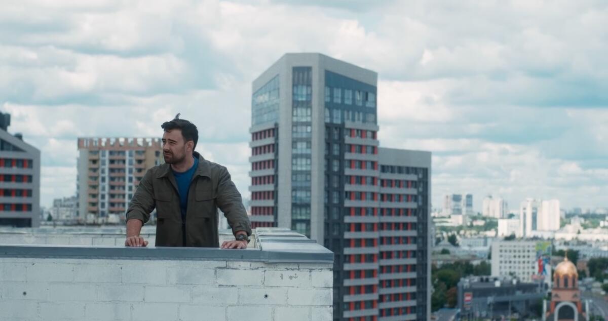 A man in a green jacket stand on a rooftop with a several tall buildings seen in the background.