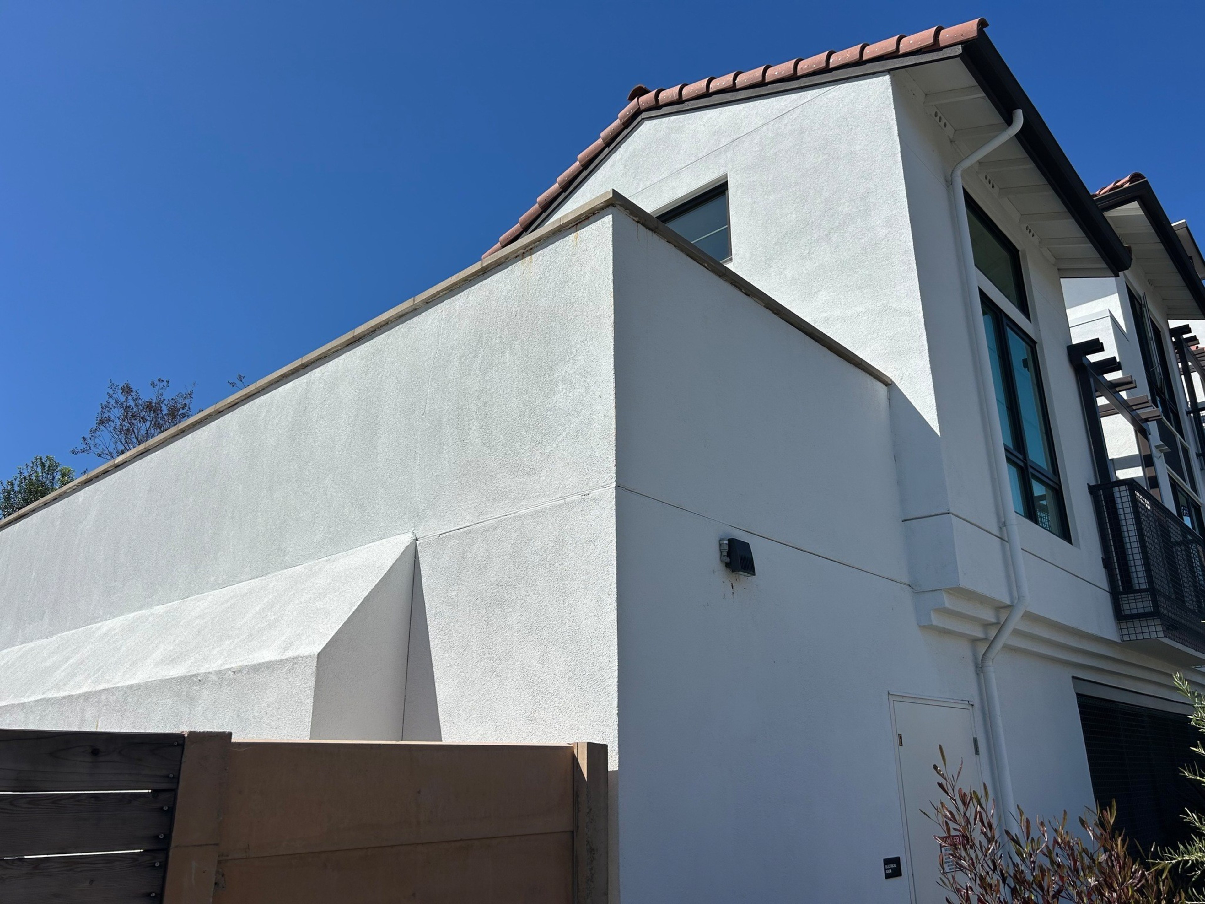 A modern white stucco building with a tile roof, large windows, a wooden gate, and a clear blue sky above.