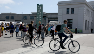 People are walking and riding bicycles near a building with a tall green sign that reads "Alcatraz Landing" on a sunny day.