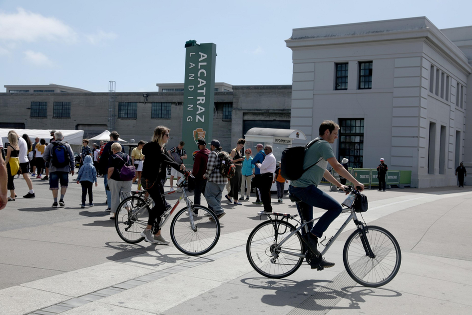 People are walking and riding bicycles near a building with a tall green sign that reads "Alcatraz Landing" on a sunny day.