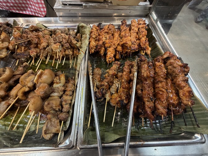 Trays of grilled meat skewers on banana leaves, including pork barbecue, chicken and offal skewers, displayed at a food service counter.