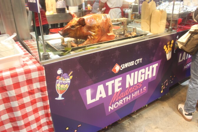 A whole roasted pig displayed on a serving counter beneath a Seafood City Late Night Madness North Hills banner.