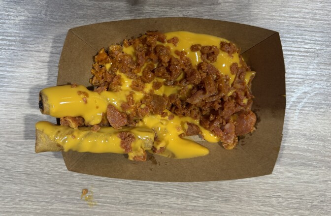 Two pieces of lumpia in a cardboard tray, topped with crumbled chorizo and drizzled with bright yellow cheese sauce.