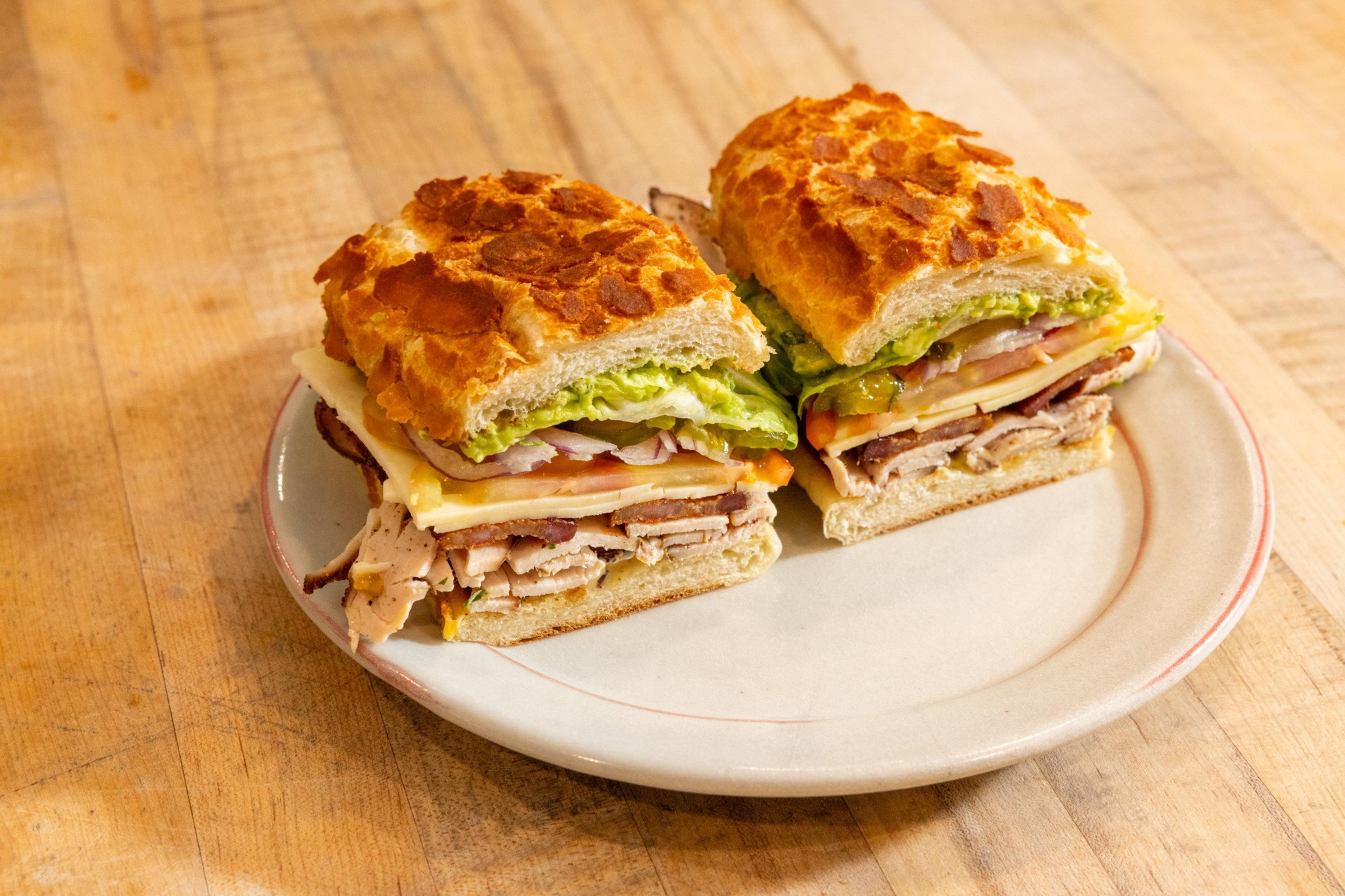 Two halves of a sandwich with lettuce, tomato, cheese, turkey, and guacamole on toasted bread sit on a white plate on a wooden surface.