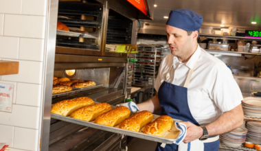 A baker wearing a blue apron and hat is removing a tray of freshly baked golden bread loaves from an industrial oven in a bakery kitchen.