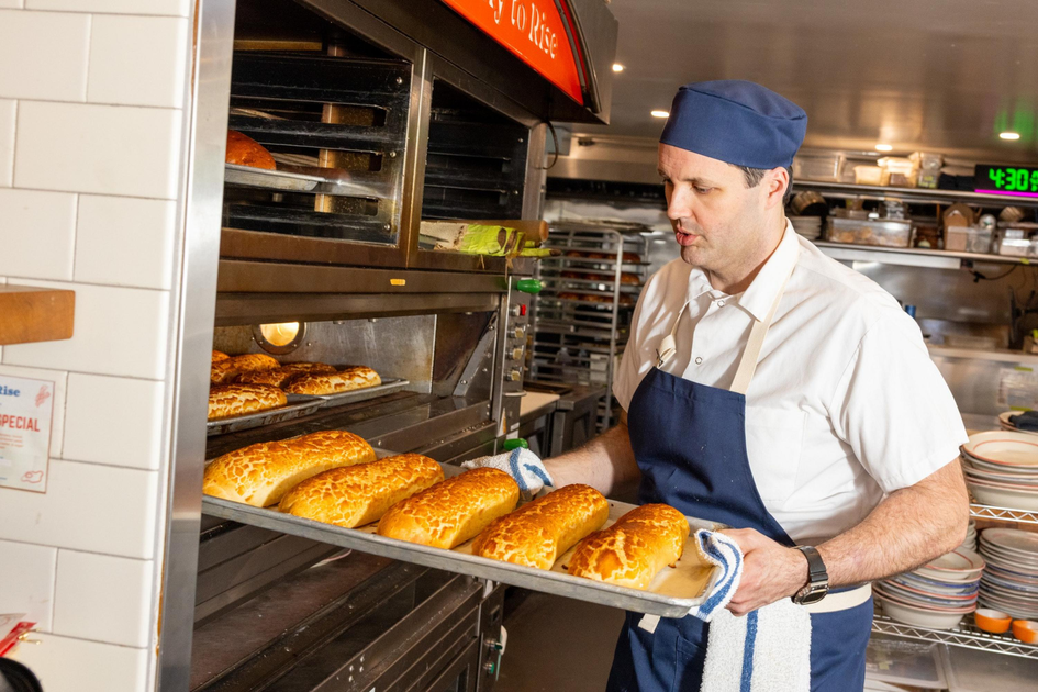 A baker wearing a blue apron and hat is removing a tray of freshly baked golden bread loaves from an industrial oven in a bakery kitchen.