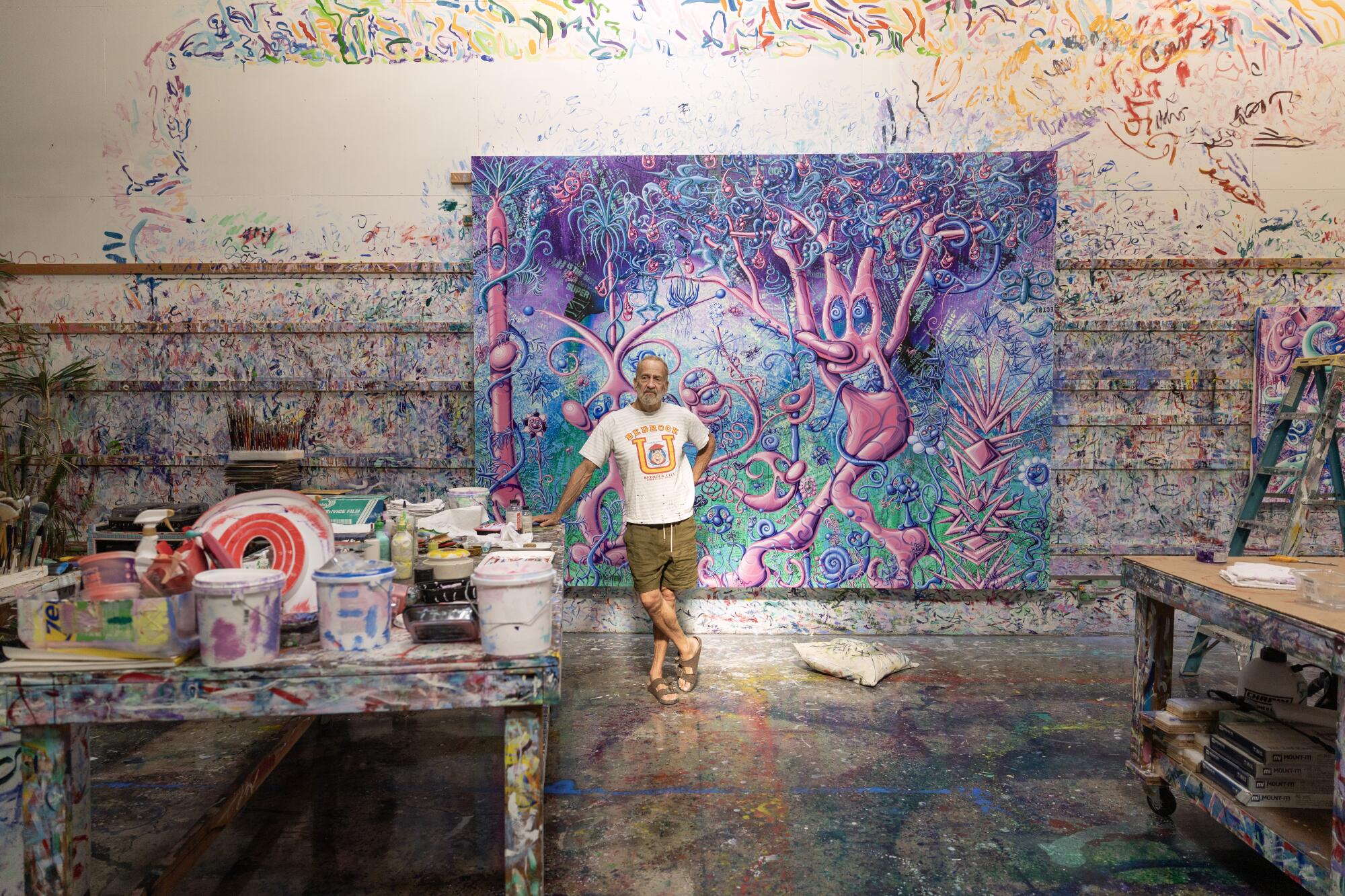 Paint covers the walls, floors, tables an a large canvas behind Kenny Scharf, wearing a T shirt and shorts.