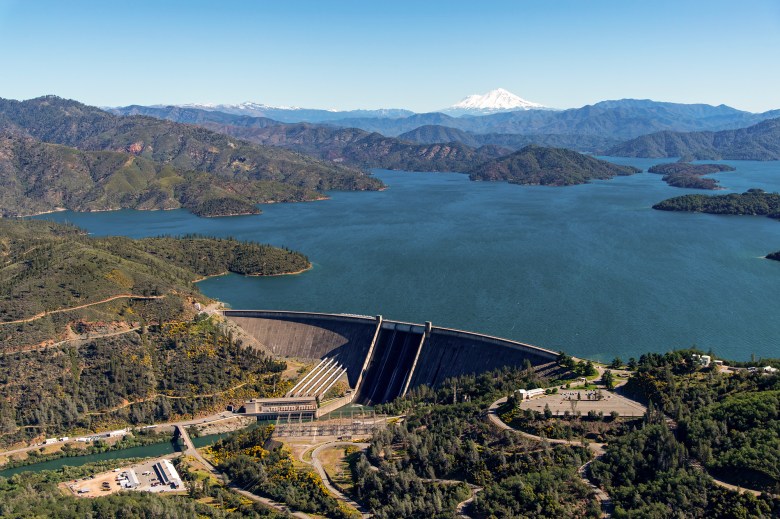 An aerial view of a large dam spanning a deep blue reservoir, surrounded by rolling green hills covered in trees and shrubs. The water extends into multiple inlets and peninsulas, creating an intricate shoreline. In the background, snow-capped mountains rise above the horizon under a clear blue sky. A hydroelectric facility and other infrastructure are visible near the base of the dam, along with winding roads leading through the landscape.