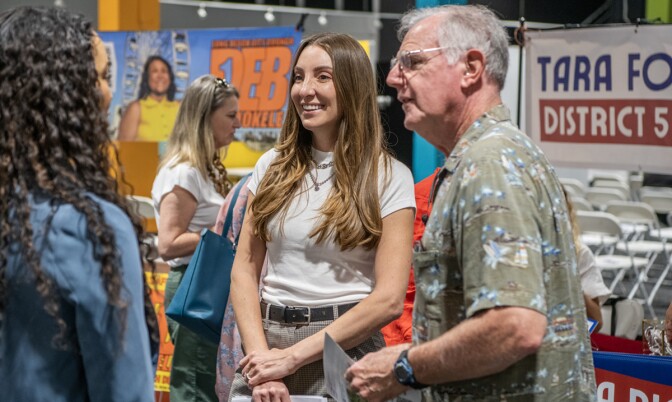 Two women, one with medium skin tone and one with light skin tone, speak with a man with light skin tone. There are people in the background talking amongst one another near white foldable chairs and banners.