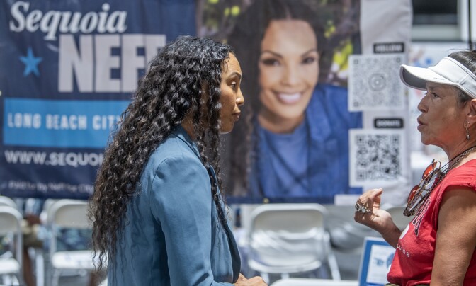 Sequoia Neff, a woman with medium skin tone, wearing an indigo coat, speaks with a woman with medium skin tone, wearing a salmon shirt. A banner hangs out of focus in the background of Sequoia Neff.