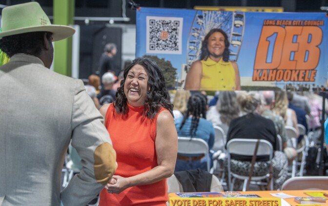 A woman with medium skin tone, wearing an orange dress, speaks with a person wearing a hat and coat. Behind her is a banner that reads "Deb Kahookele" with an image of her and people sitting in foldable white chairs facing the other direction.
