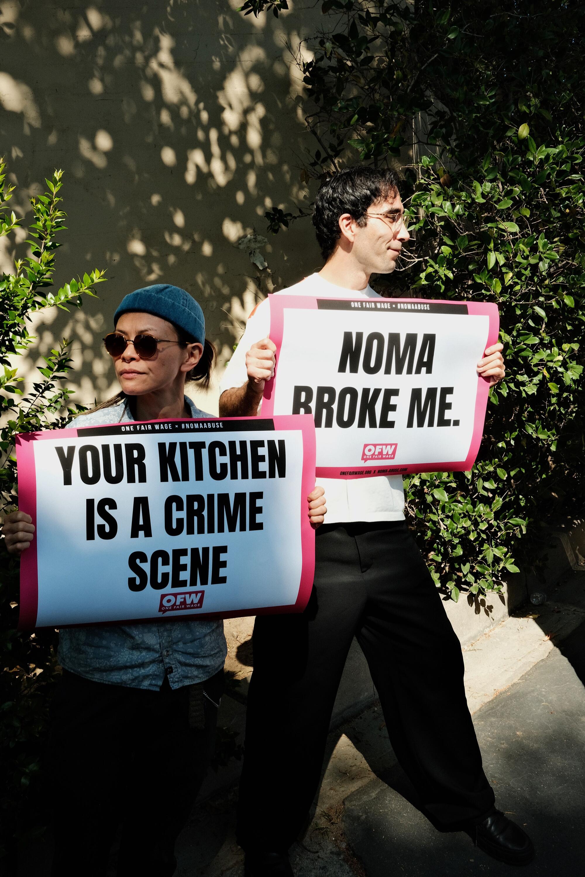 Two people stand holding pink, black and white protest signs that say "Noma broke me" and "Your kitchen is a crime scene"