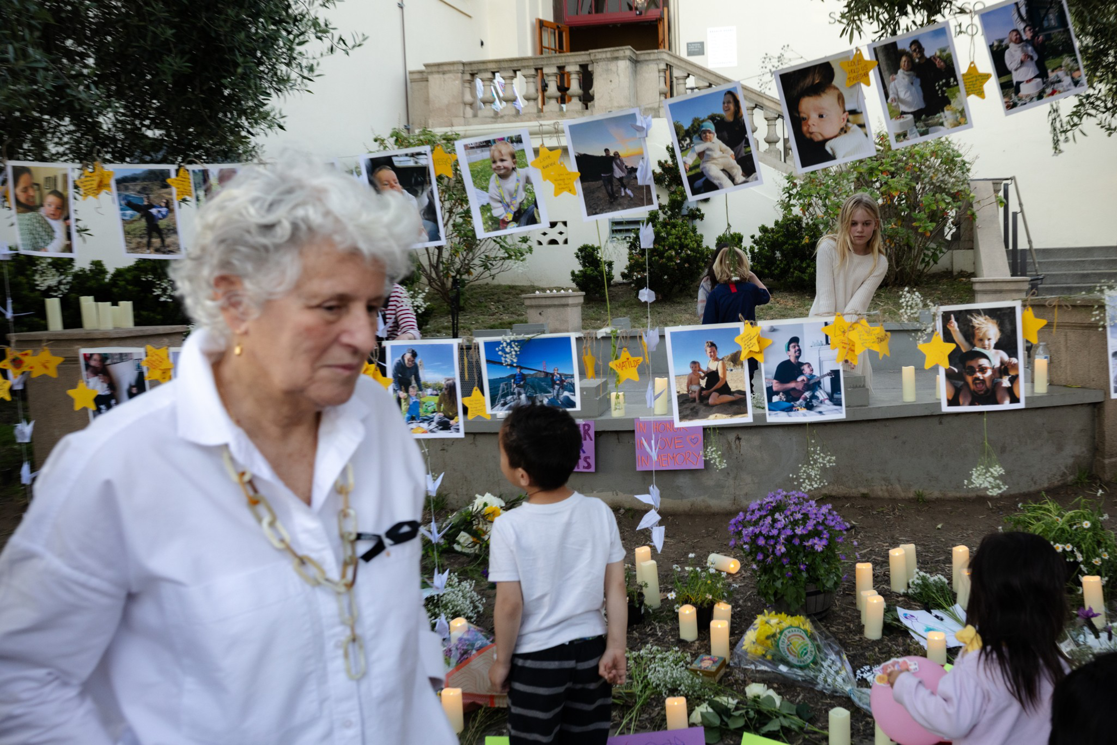 Photos of various people are displayed on strings with yellow star decorations, surrounded by flowers and candles, while people observe quietly nearby.