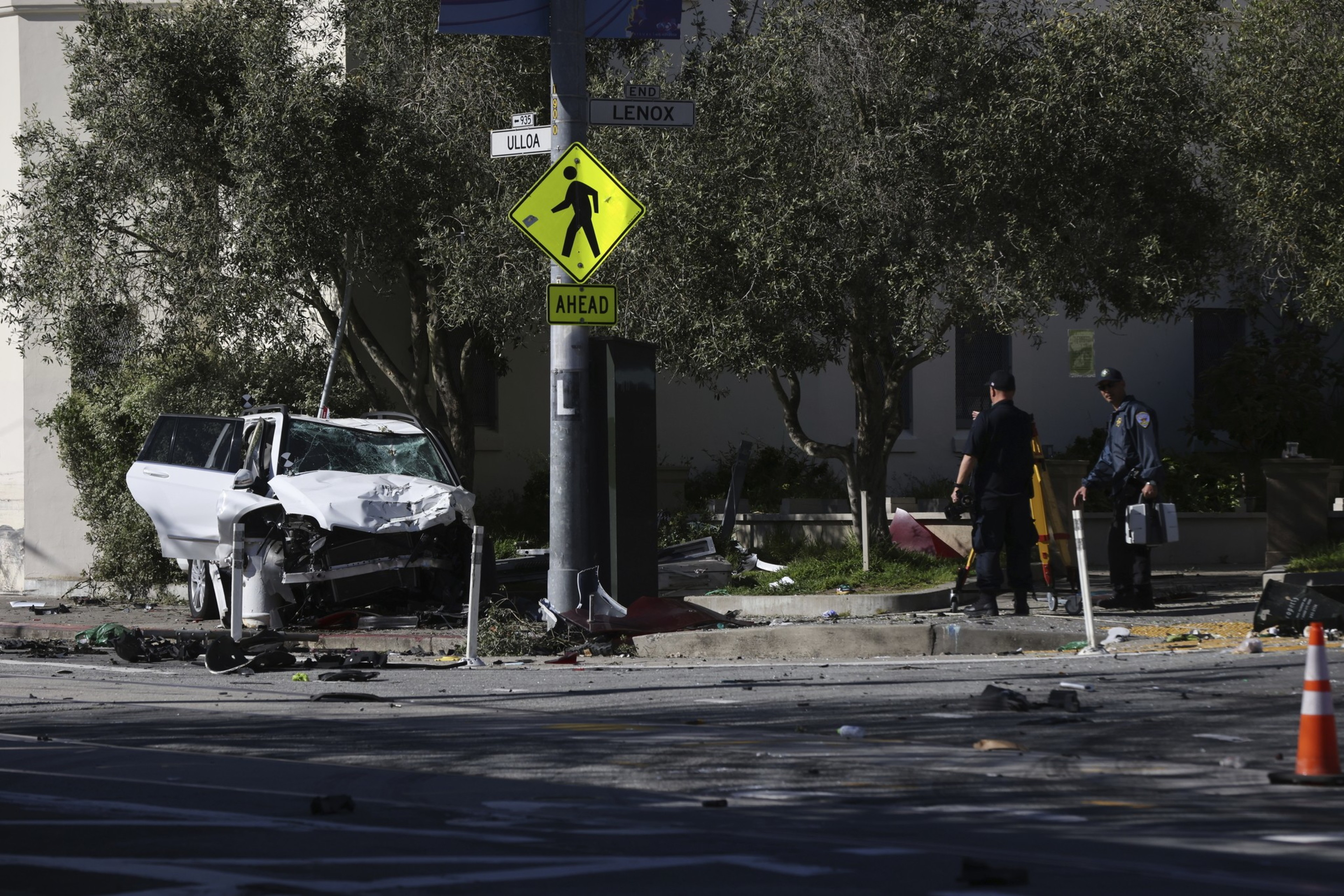A damaged white car is crashed into a pole near a pedestrian sign. Debris litters the street, and two officers investigate the scene with caution tape around.
