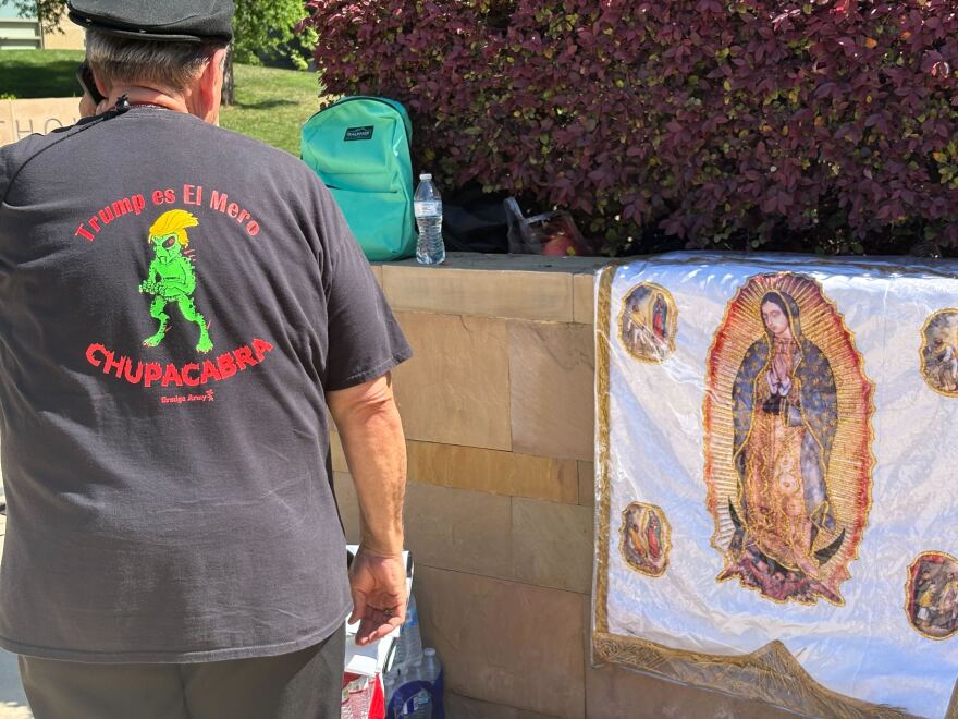 Image of a rally participant wearing a T-shirt printed with an alien and the message "Trump is the Ultimate Sucker"—in Spanish—standing in front of a flag of the Virgin of Guadalupe on Wednesday, March 18, 2026.