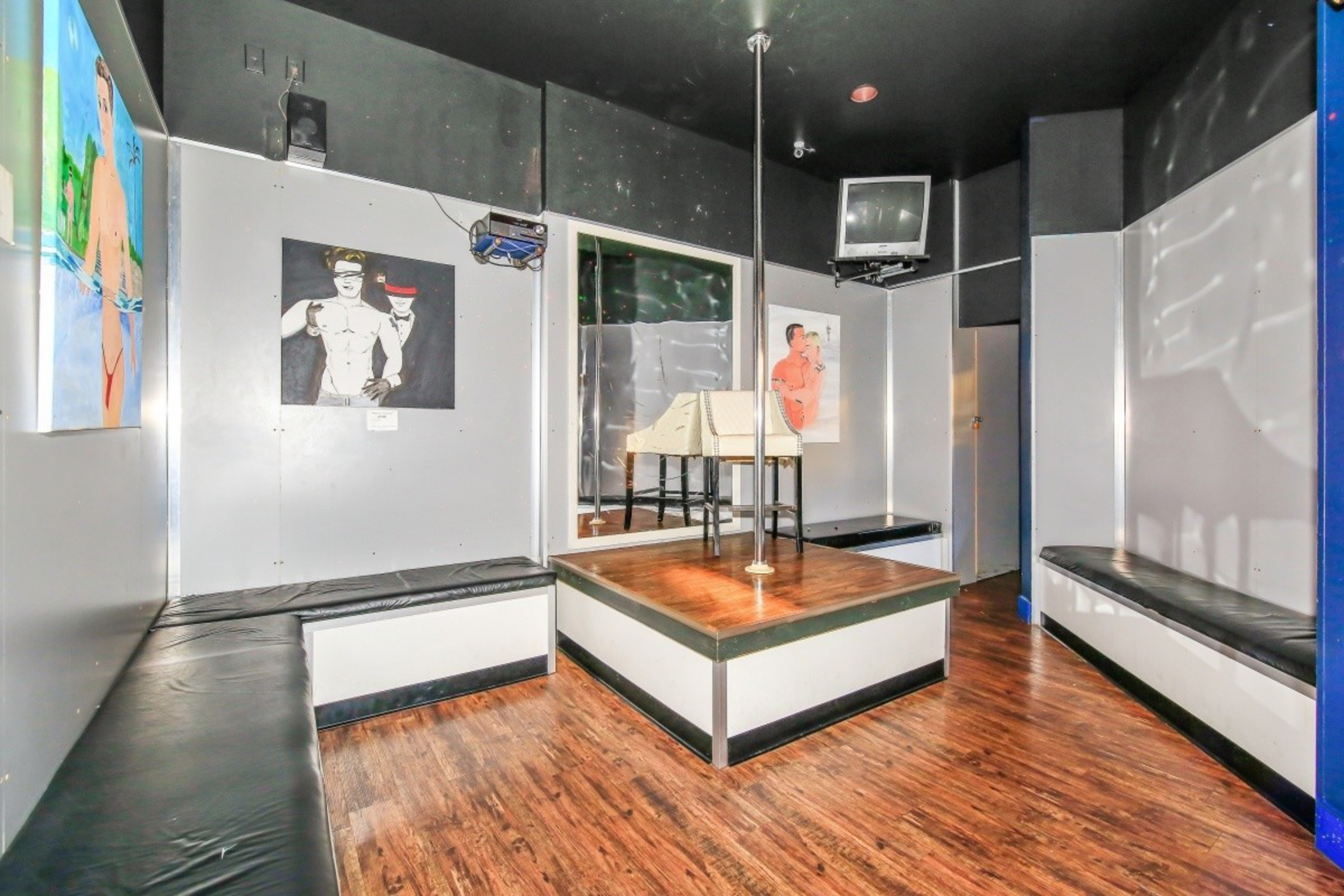 A small room with a wooden stage and pole in the center, black cushioned benches on both sides, three paintings of shirtless men, a wall mirror, and a mounted TV.