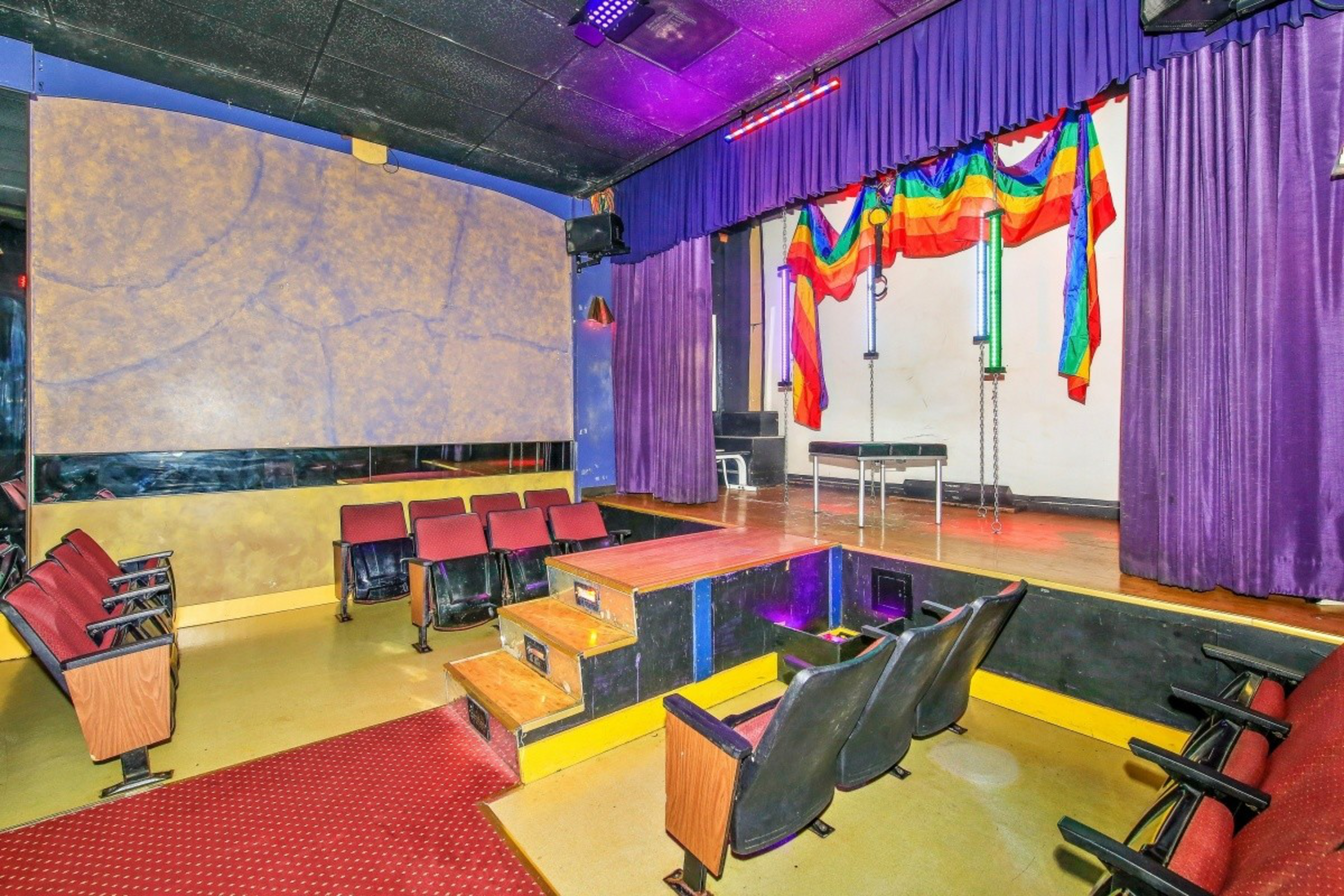 A small theater room with red cushioned seats, a yellow floor, a stage with rainbow flags draped over purple curtains, and a red carpeted aisle.