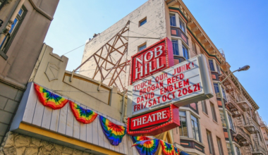 Nob Hill Theatre marquee displays upcoming shows with rainbow decorations along a stone wall beneath a clear blue sky.