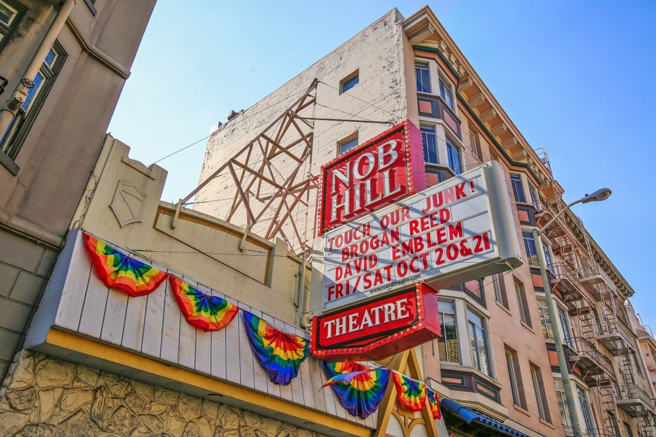 Nob Hill Theatre marquee displays upcoming shows with rainbow decorations along a stone wall beneath a clear blue sky.