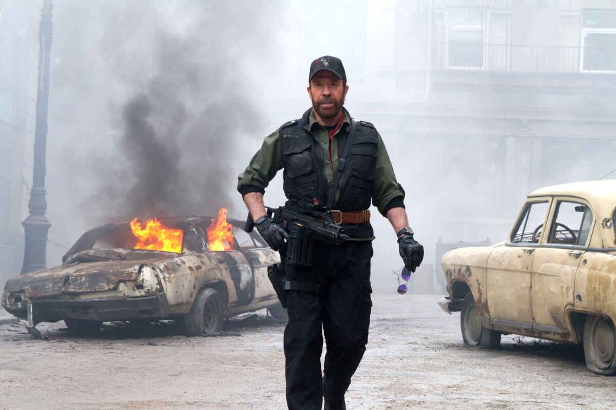 Chuck Norris in character walking between wrecked cars, one of them on fire, in "The Expendables 2"