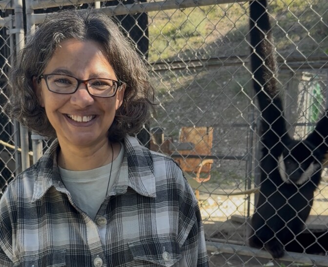 A woman with black curly hair smiles. There is a black gibbon primate in a cage behind her. 