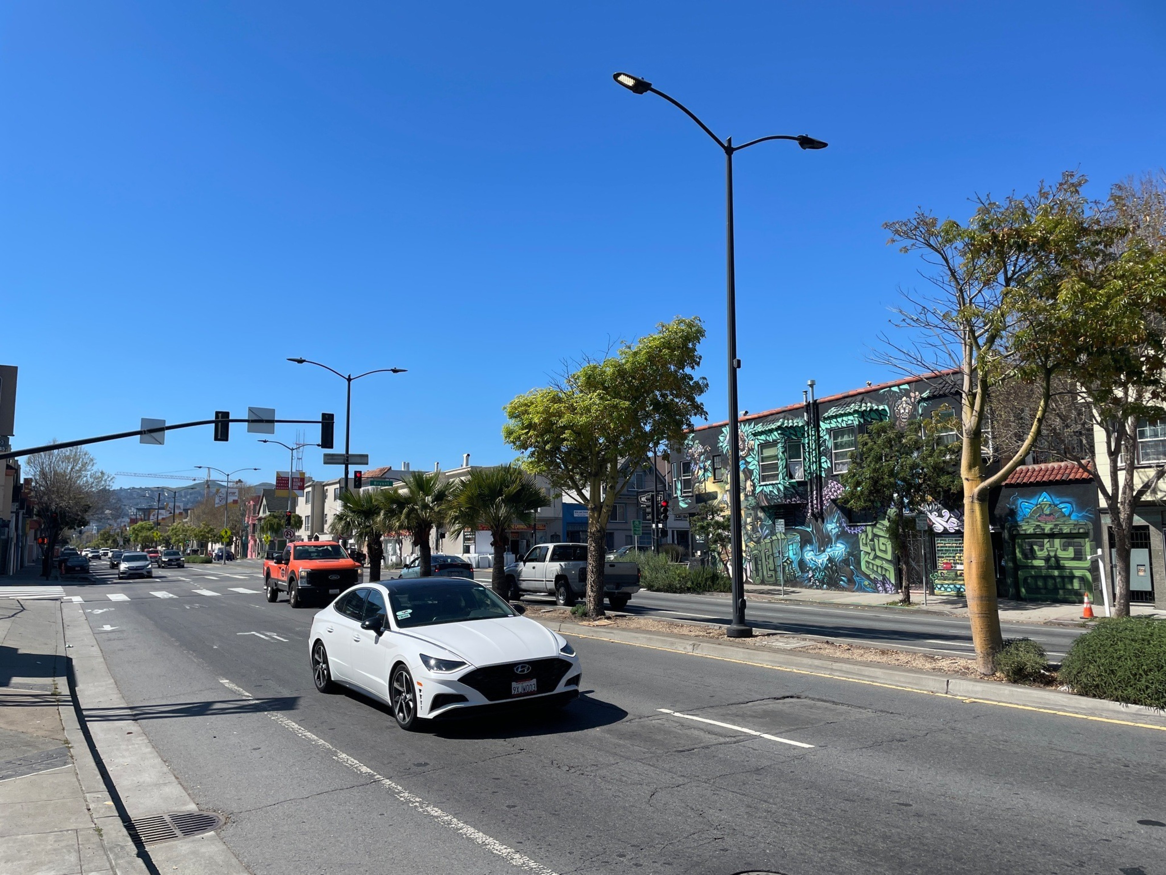 A white car drives on a sunny street lined with palm trees, colorful building murals, and light traffic under a clear blue sky.
