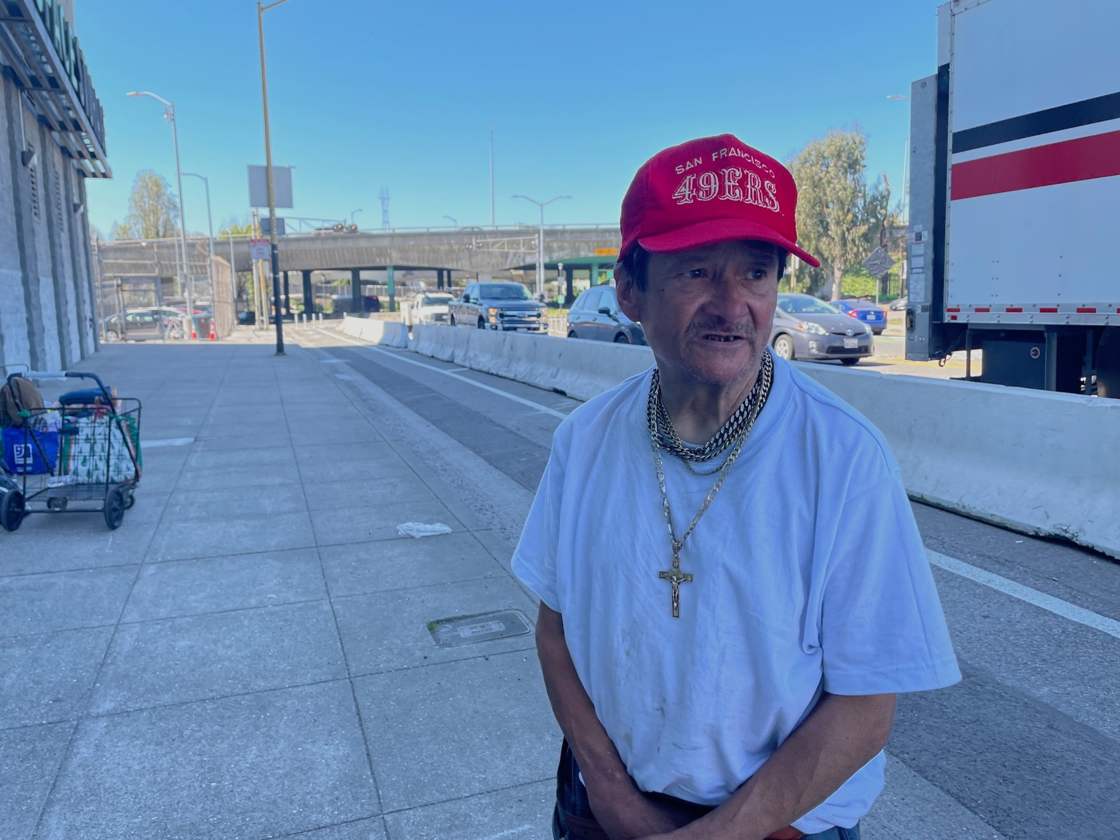 A man in a red San Francisco 49ers cap and white shirt with a gold cross necklace stands on a sidewalk near a busy street with cars and a truck.