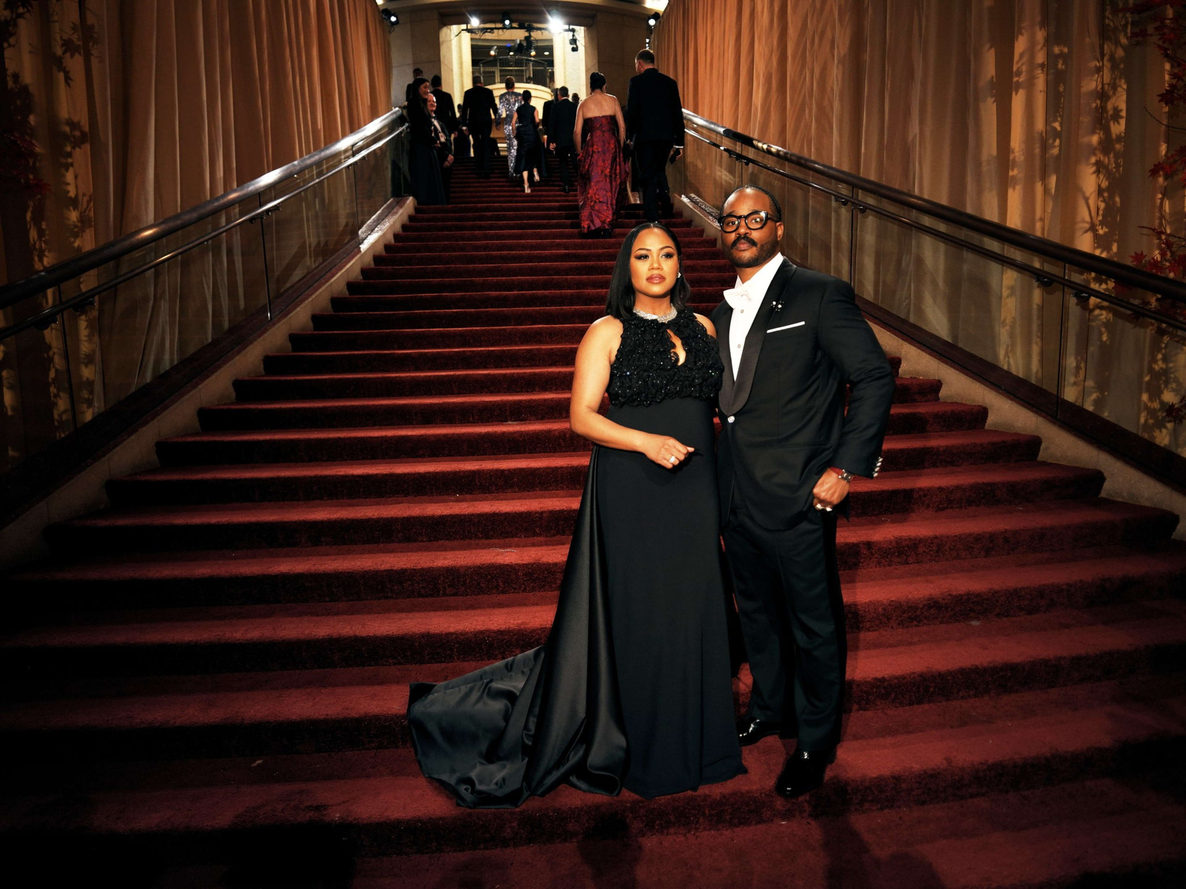 A man in a black tuxedo and a woman in a long black gown stand side by side at the bottom of a grand, carpeted staircase with gold handrails.