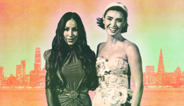 A woman with wavy, light brown hair wears tortoiseshell glasses, bright pink lipstick, a black blazer, and a black and white patterned high-neck top.