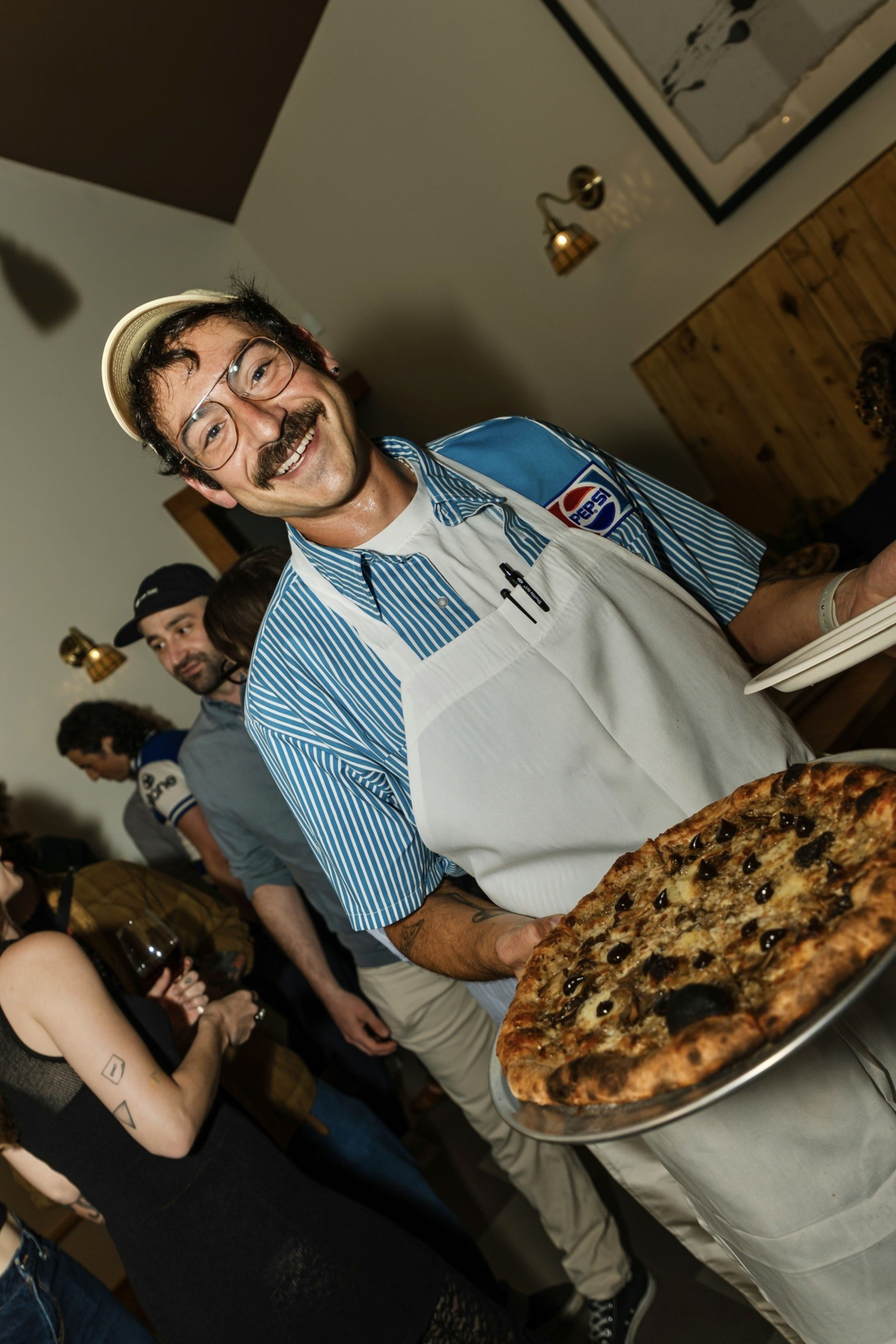 A smiling man with glasses, a mustache, and a striped shirt holds a large pizza while wearing a white apron in a crowded indoor setting.