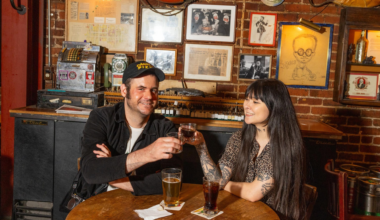 A man and woman sit at a wooden table in a cozy bar, smiling and clinking small glasses with drinks, surrounded by vintage decor and framed pictures.
