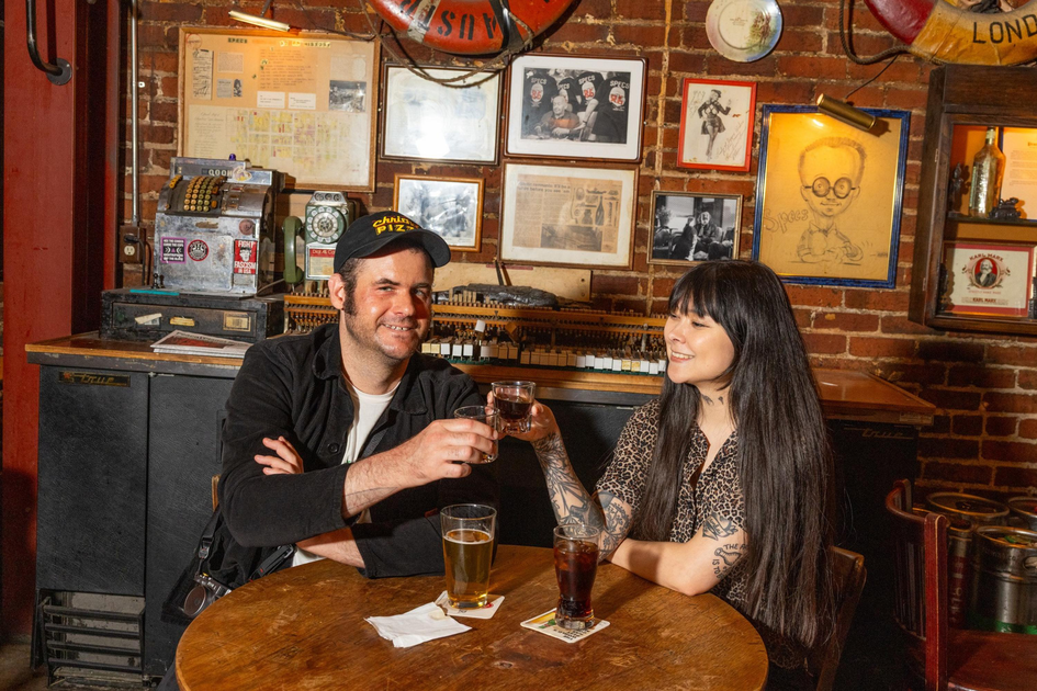 A man and woman sit at a wooden table in a cozy bar, smiling and clinking small glasses with drinks, surrounded by vintage decor and framed pictures.