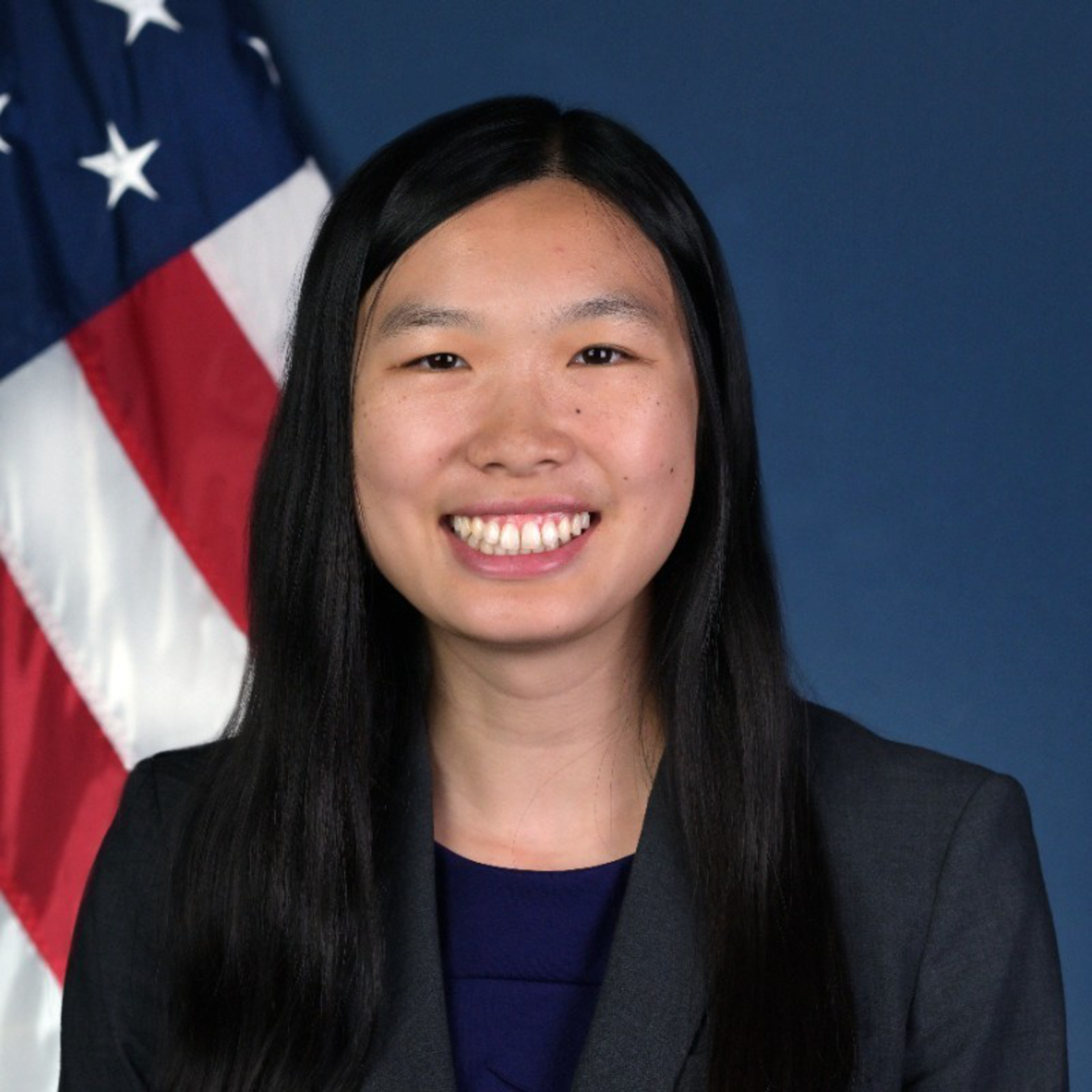 A smiling woman with long black hair wears a dark blazer over a blue top, standing against a blue background with a US flag visible behind her.