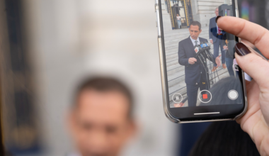 A person is recording a man in a suit speaking at a microphone outside a building, with the video clearly visible on the phone screen.