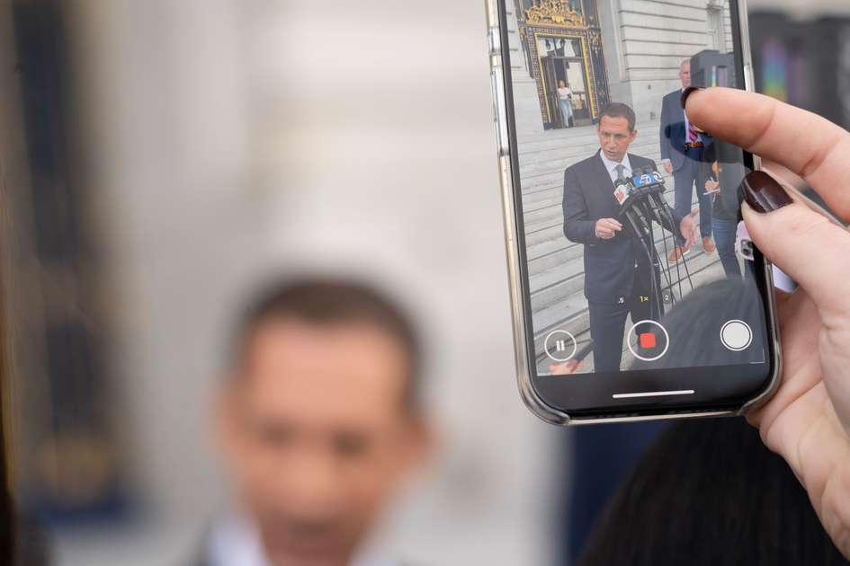 A person is recording a man in a suit speaking at a microphone outside a building, with the video clearly visible on the phone screen.