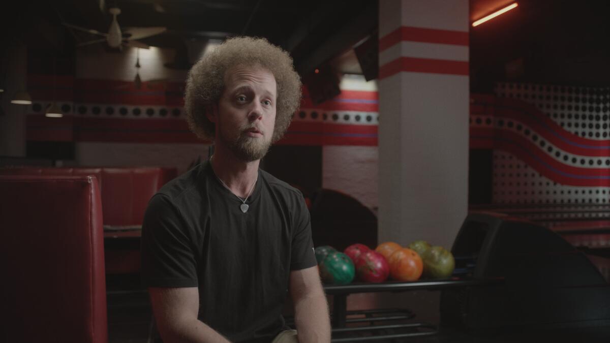 A man in black T-shirt sits in a bowling alley.
