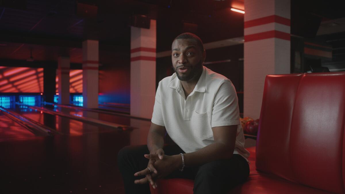 A man in a white shirt and dark slacks sits on a red bench in a darkly lit bowling alley.