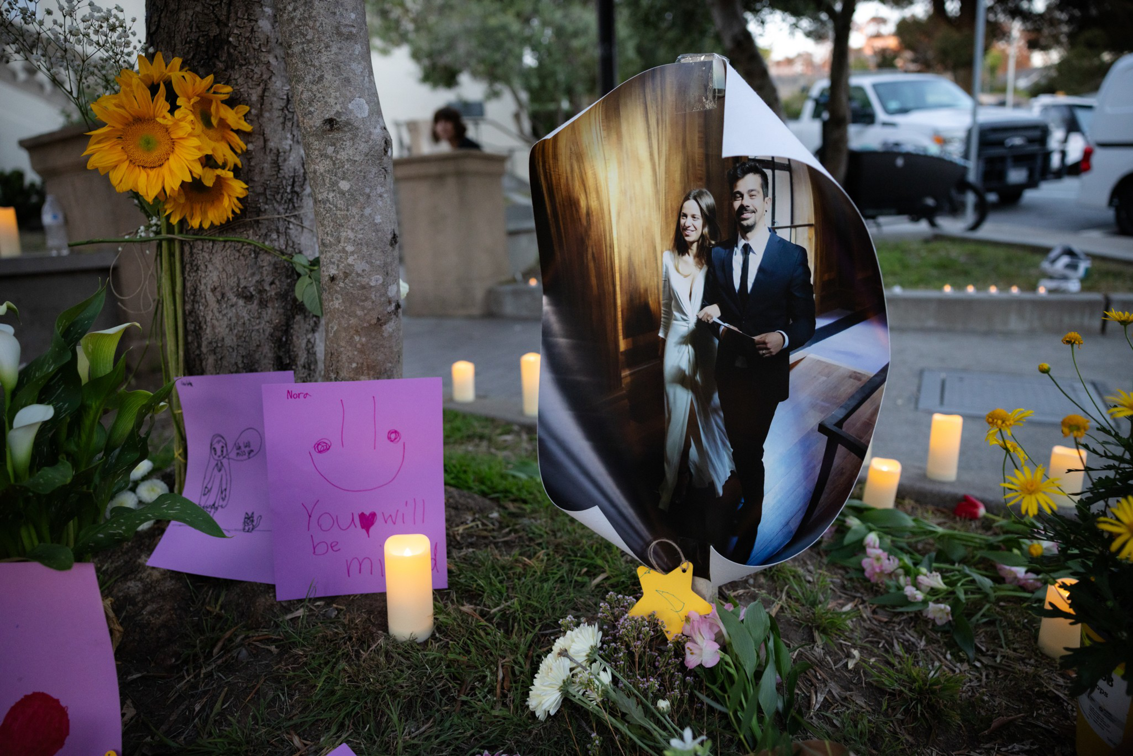 A memorial with candles, flowers, children’s drawings, and a large photo of a smiling couple dressed formally, placed outdoors near a tree.