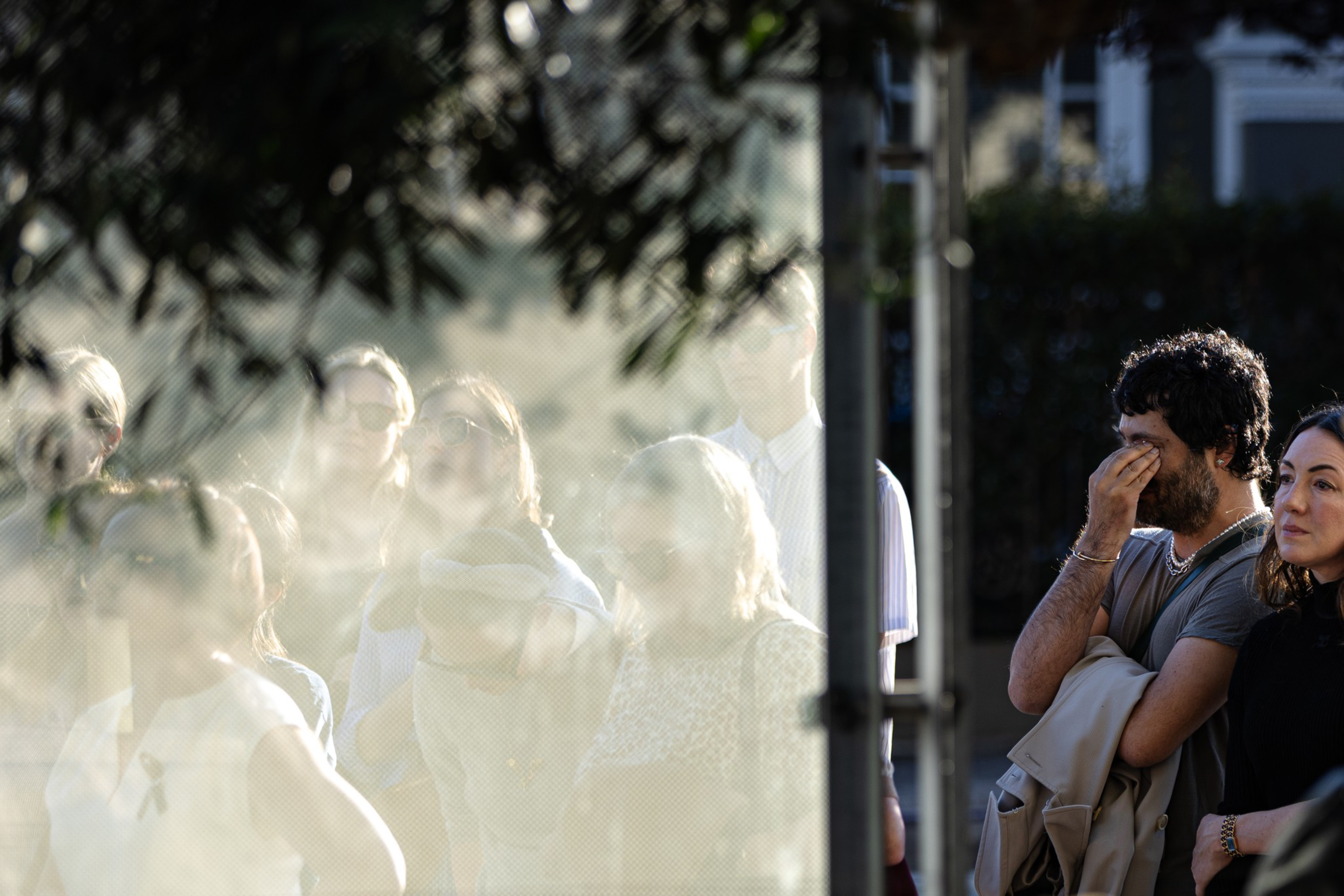 A group of people stands quietly behind a translucent screen, with two individuals on the right side in sharp focus, one covering his face.