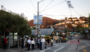 People wait at a street corner near a tram stop with overhead wires, surrounded by trees and buildings on a hill during late afternoon.