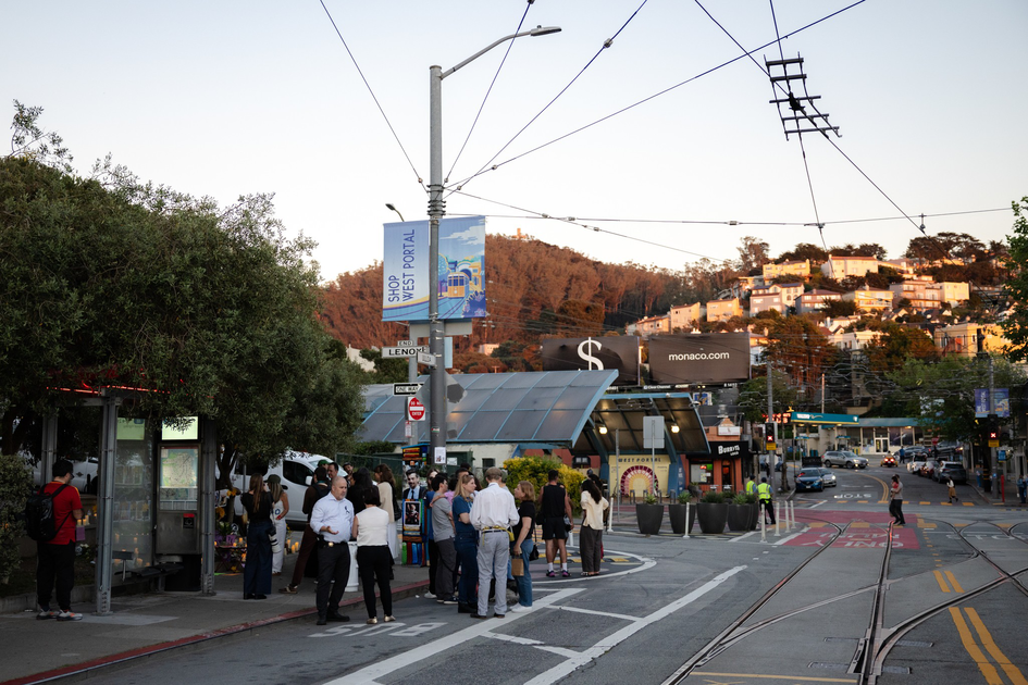 People wait at a street corner near a tram stop with overhead wires, surrounded by trees and buildings on a hill during late afternoon.