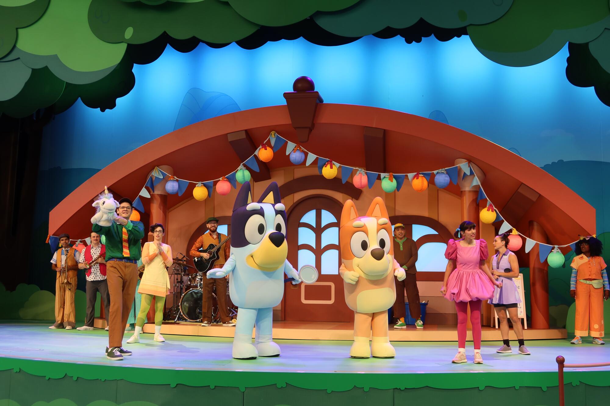 "Bluey's Best Day Ever!" cast with pastel-colored costume puppies stands on a stage in front of a house exterior