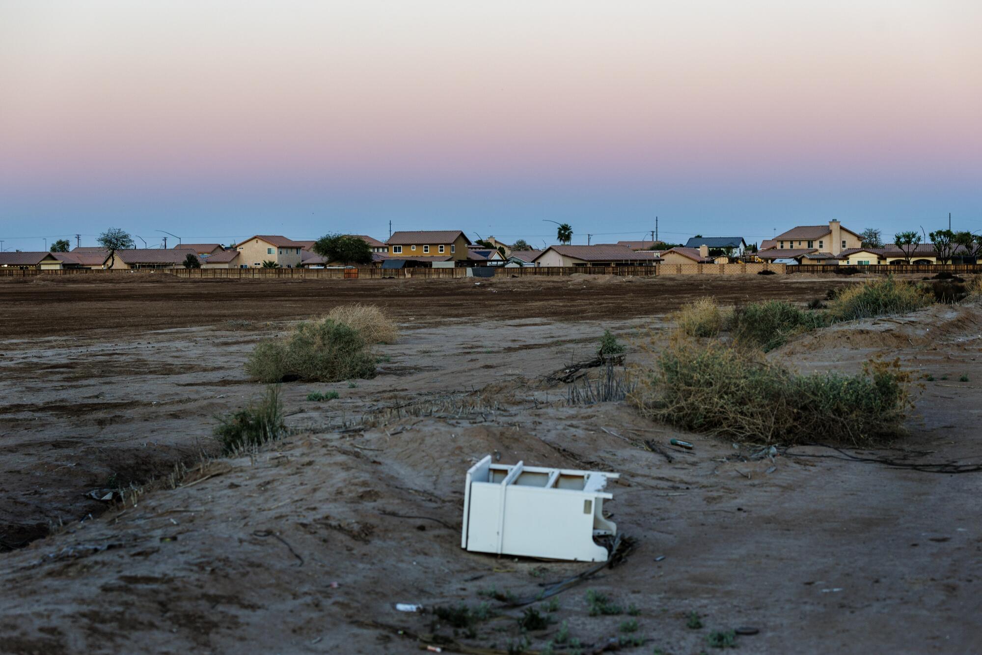 A cabinet rests on its side in the dirt on open land with houses and sky in the background.