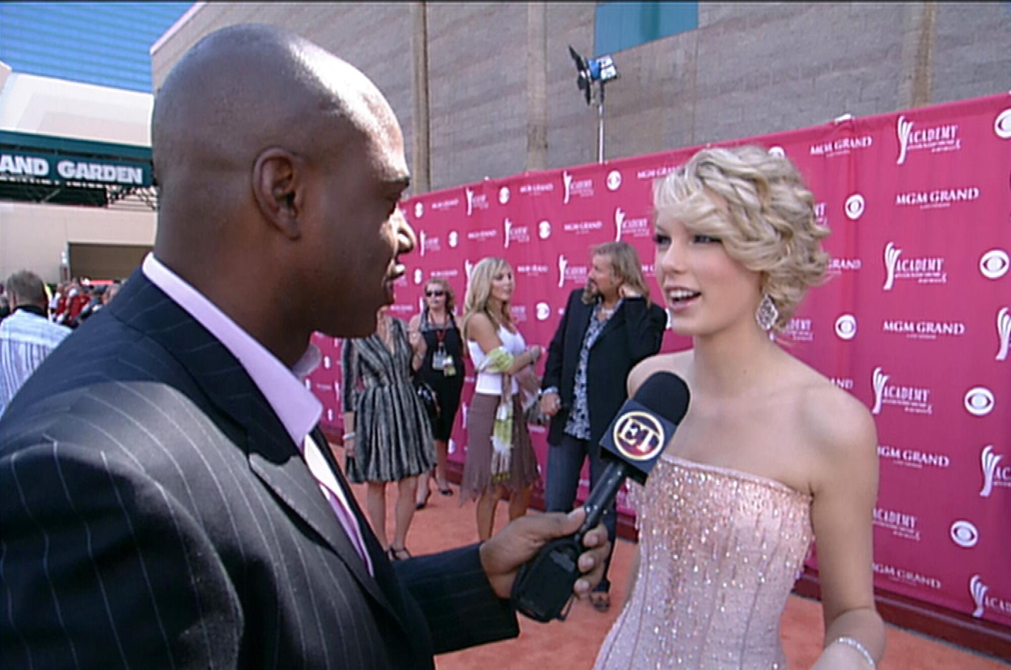 A man in a black pinstripe blazer holds a microphone toward a teenage girl in a pink sparkly strapless gown.