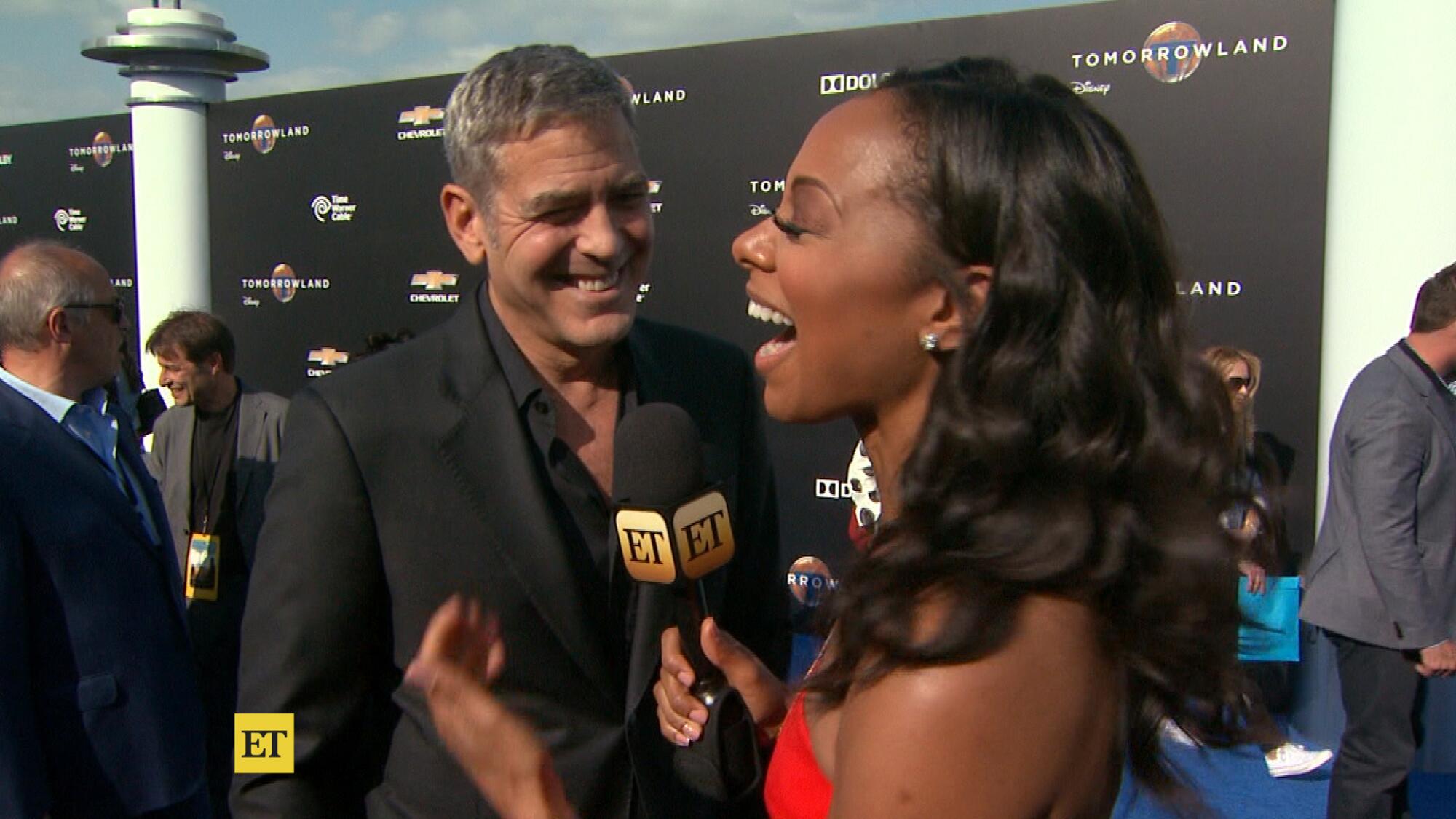 A man in a black suit and shirt speaks to a woman in a red dress holding a microphone and seemingly laughing.