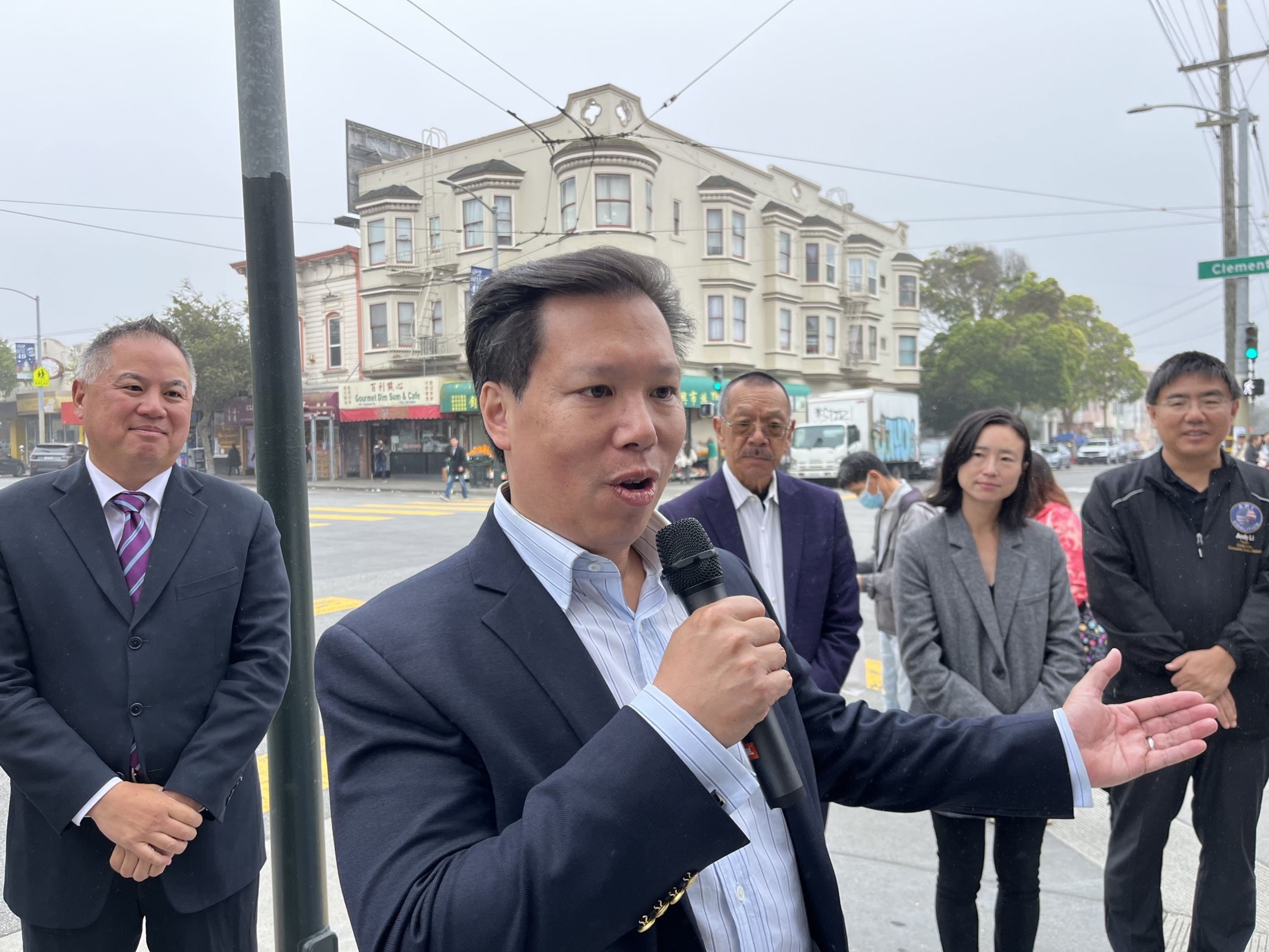 A man speaks into a microphone on a city street, with four onlookers behind him, building and foggy sky in the background.