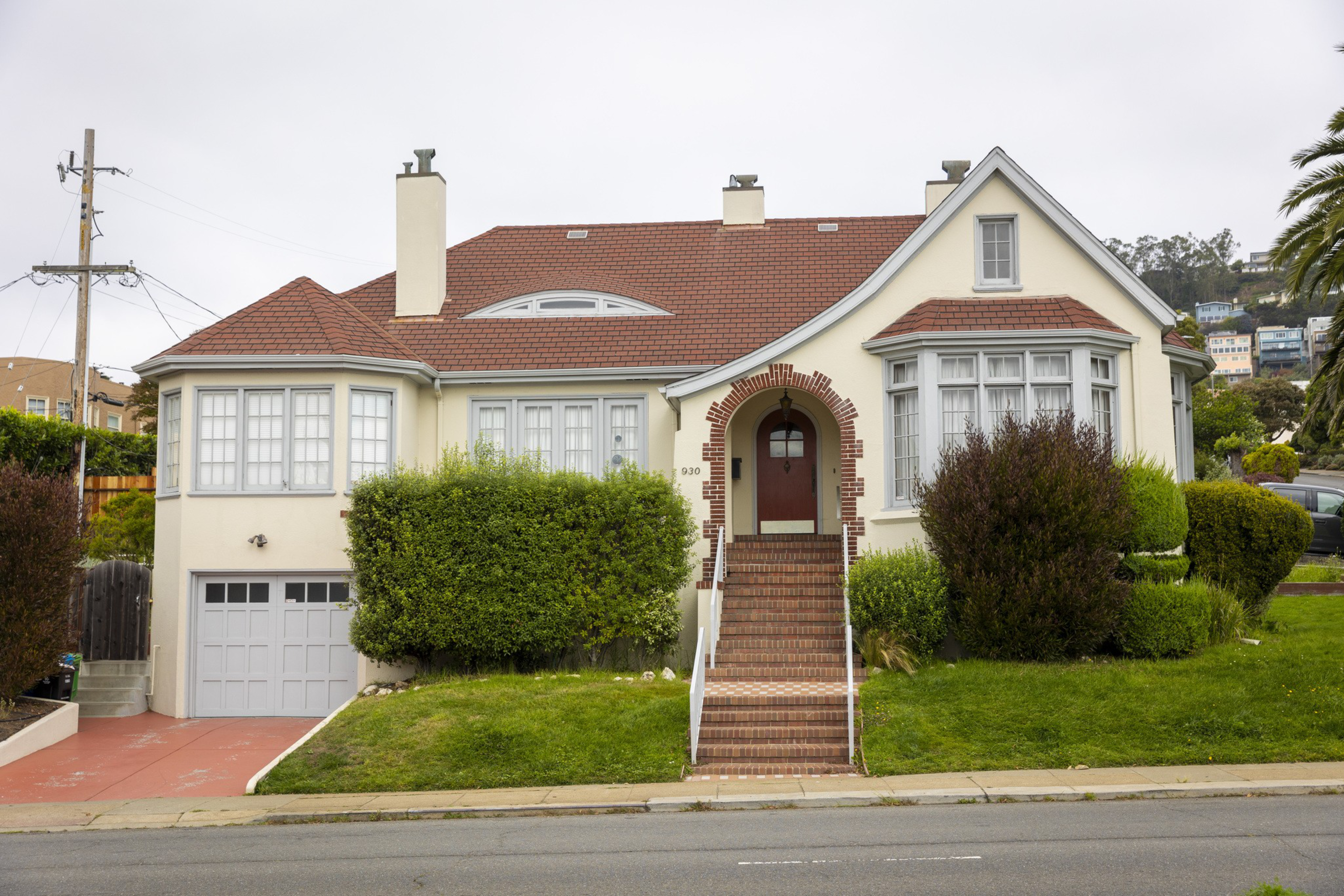 A cream-colored house with a red-tiled roof, brick steps leading to a red front door, large windows, a garage on the left, and trimmed bushes in front.