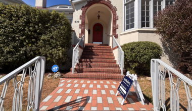 A brick staircase with white railings leads to a red door under an arch with a hanging light, flanked by bushes and a Sotheby’s real estate sign.